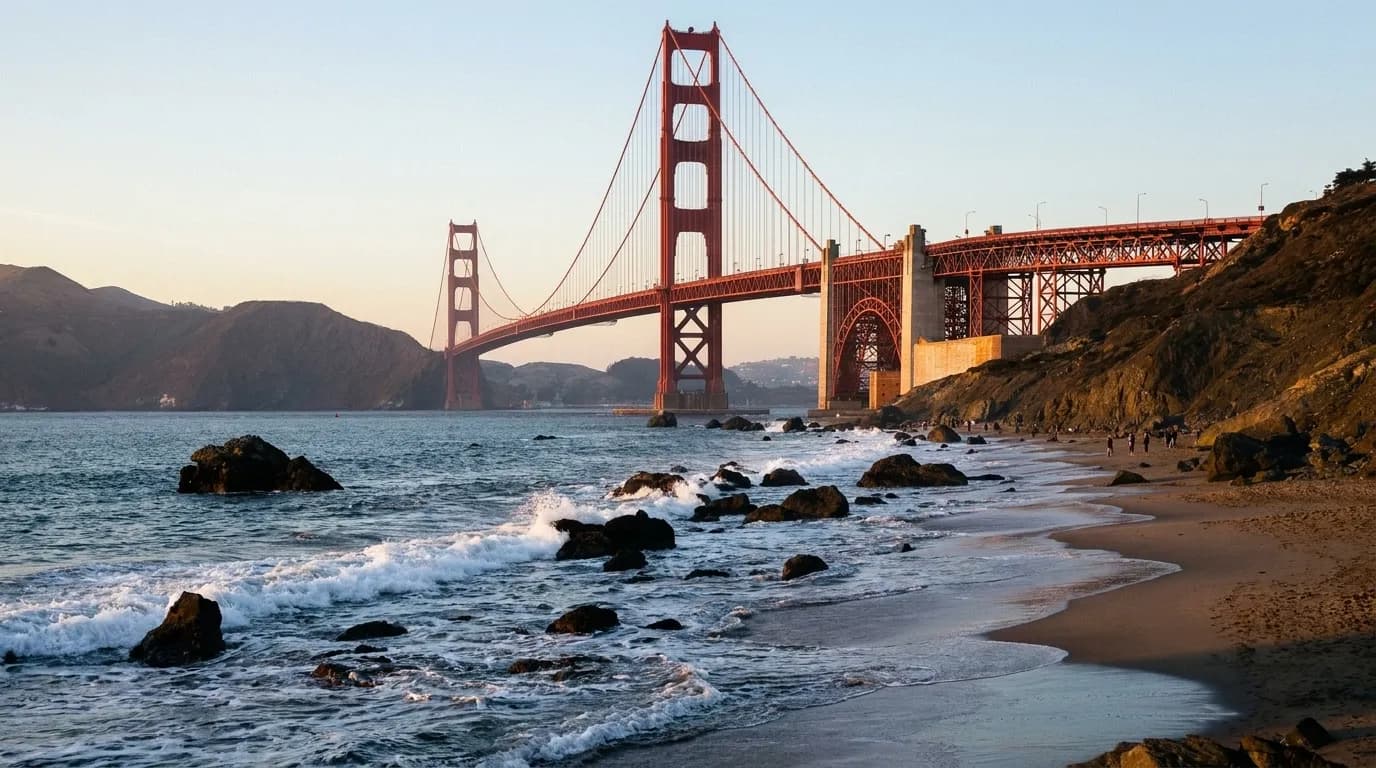 A wide San Francisco beach in late afternoon with cliffs at one end, the Golden Gate Bridge towers visible through haze and a scatter of people on the sand