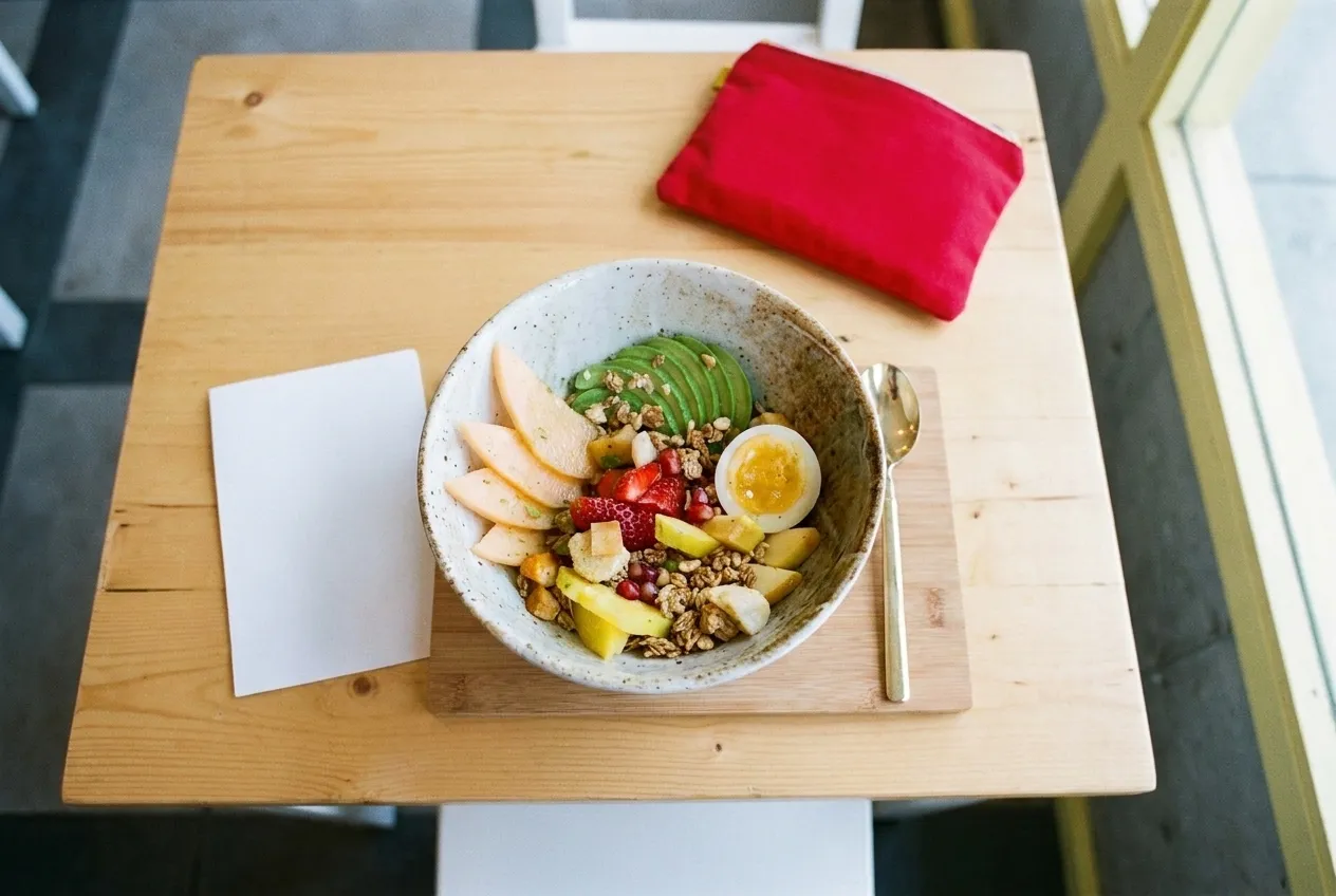 A bowl of fruit and granola on a pale table seen from directly above