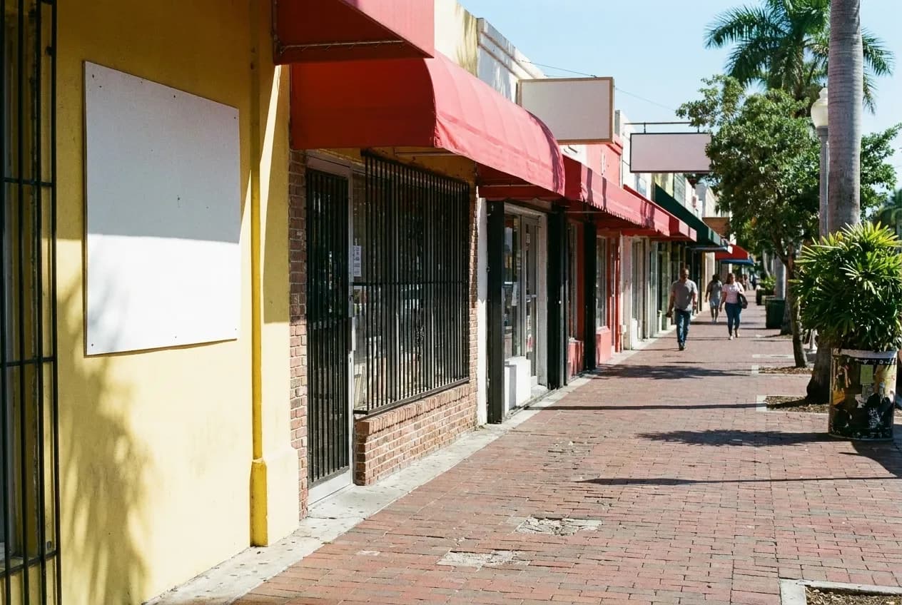A sunlit pavement running past a row of small shopfronts with awnings and barred windows, a hand-painted sign panel left blank on one wall
