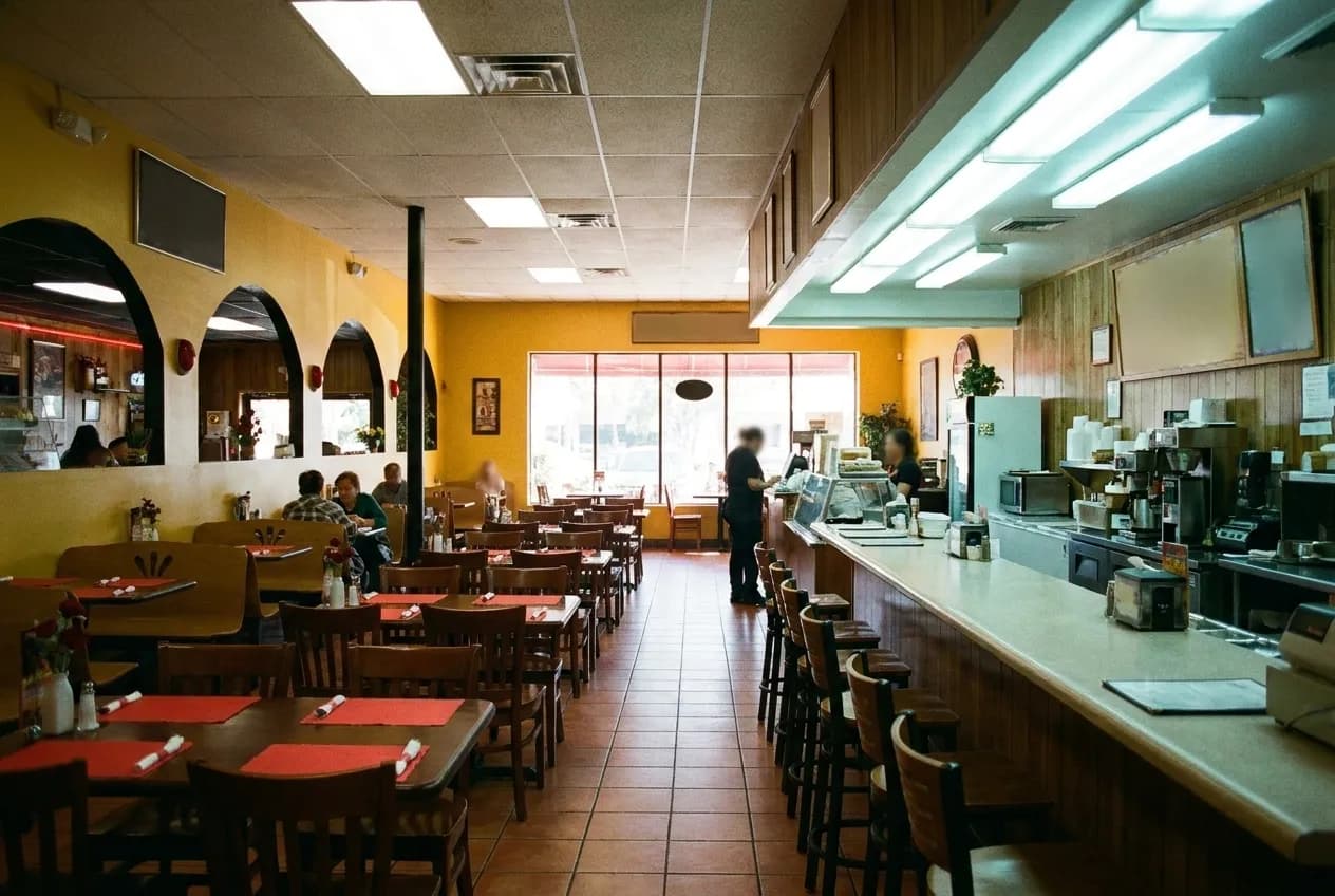 A bright restaurant interior with closely spaced tables, tiled floor and a long service counter running down one side under fluorescent light