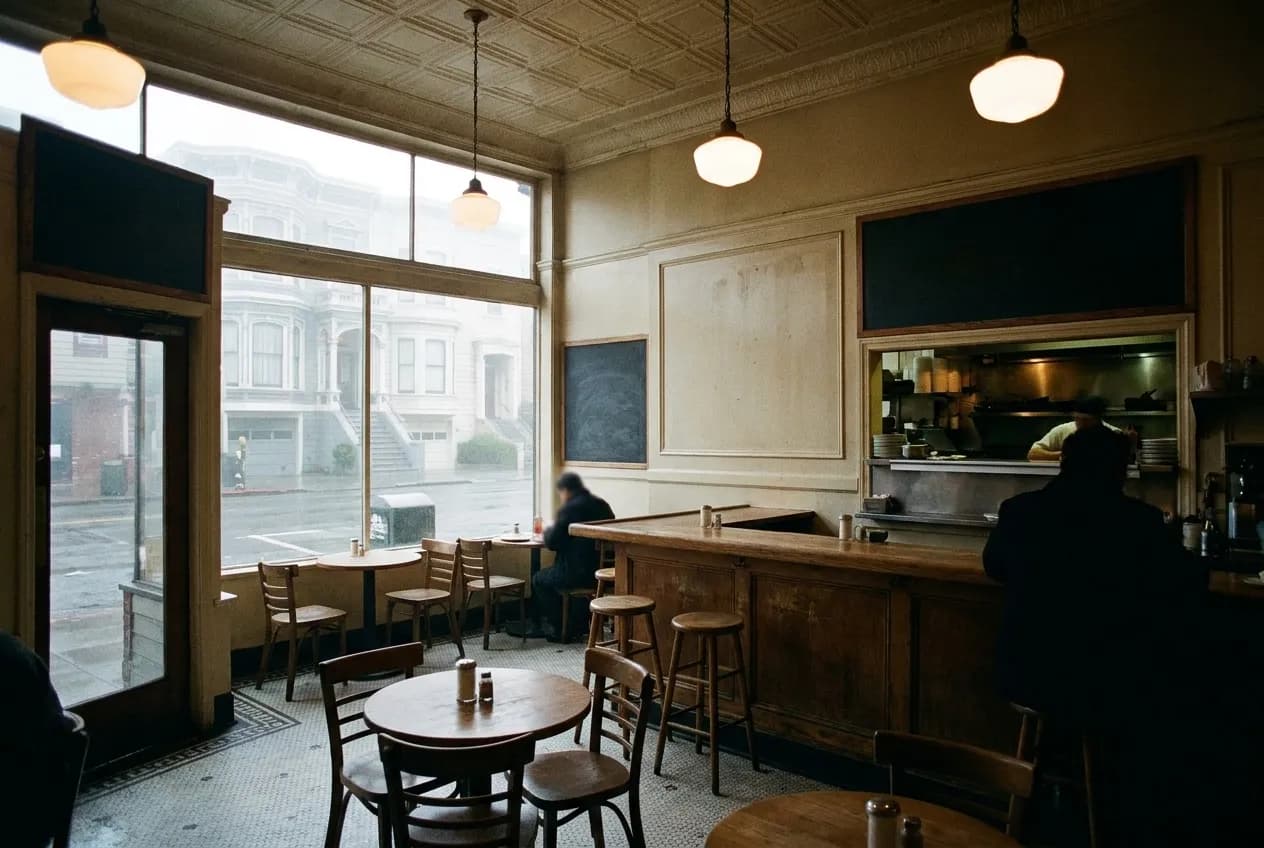 A modest restaurant interior with a short counter, a handful of tables and pendant lights, tiled floor and a serving hatch to the kitchen