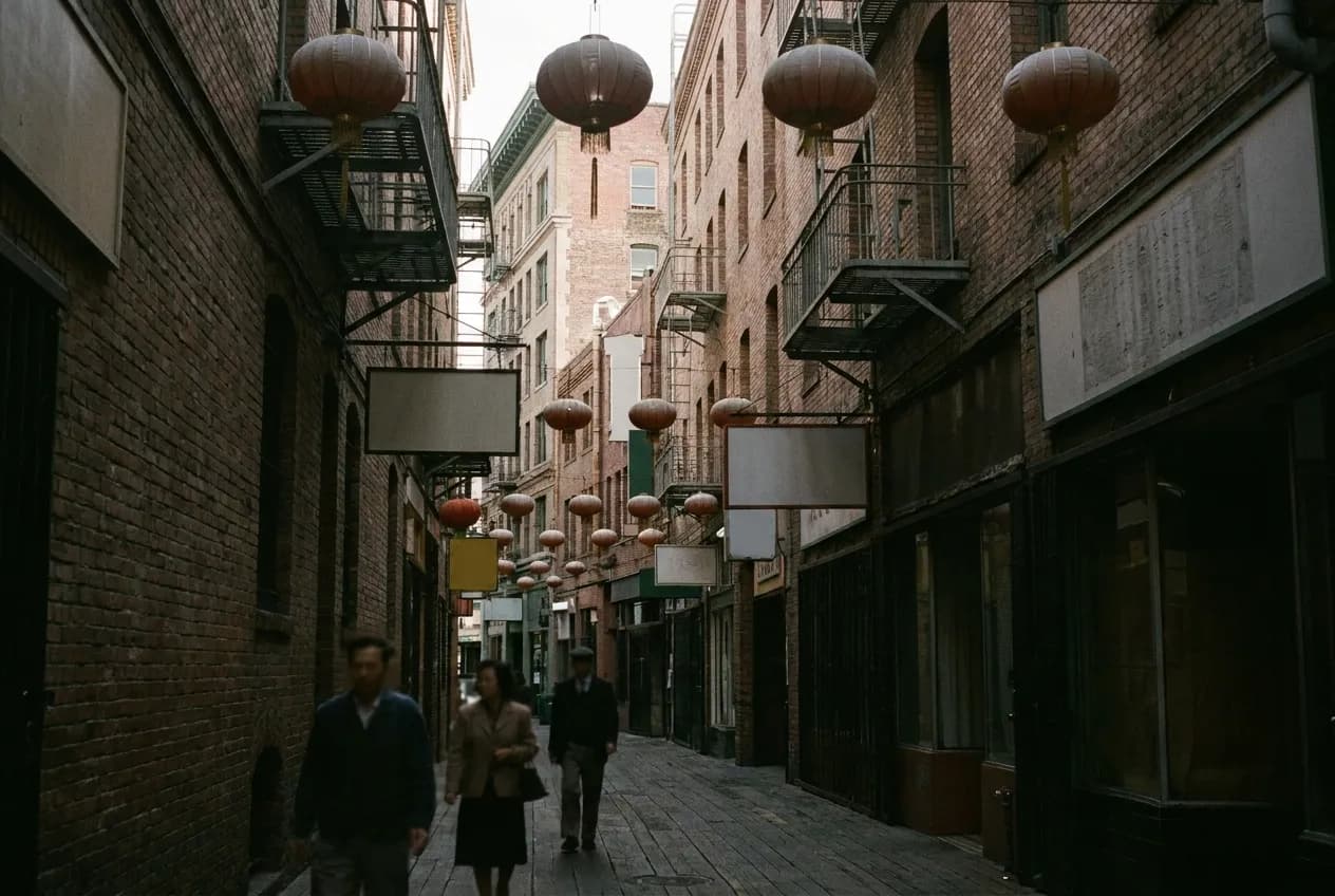 A narrow alley between tall brick buildings hung with lanterns and fire escapes, daylight reaching only the upper walls