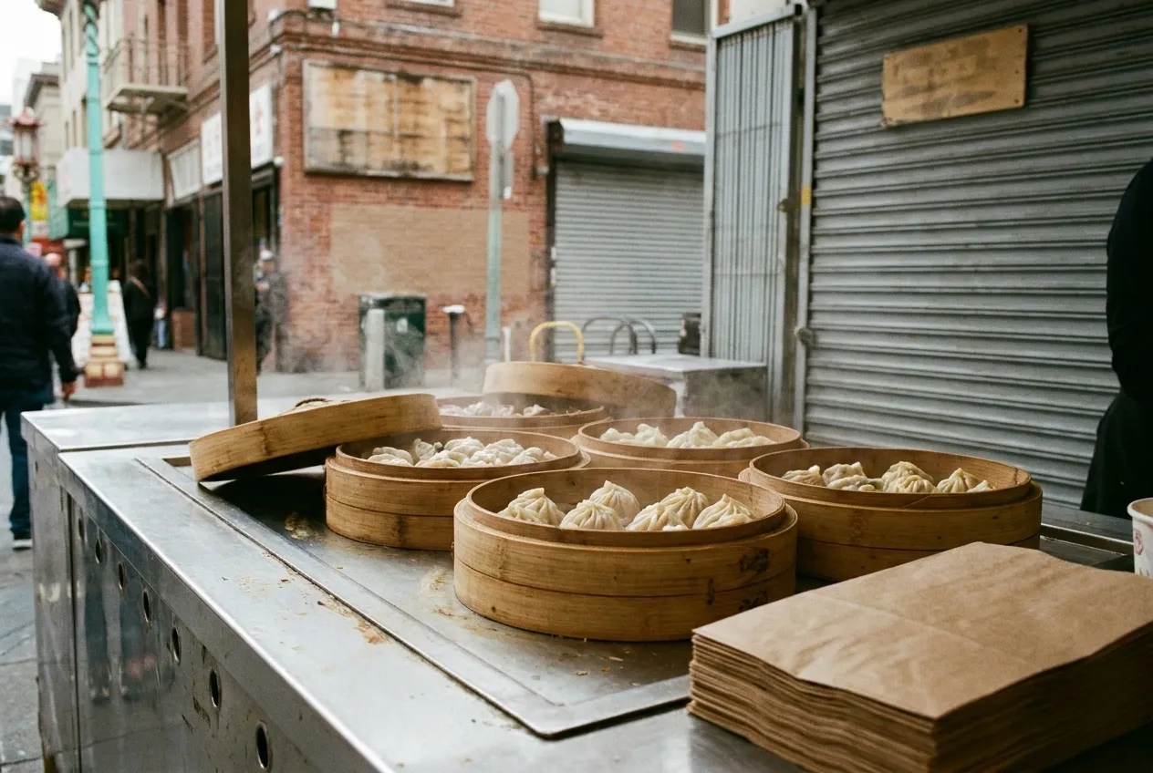 Open bamboo steamer baskets of pleated dumplings on a stainless counter, steam still rising and a stack of paper bags at the edge of frame