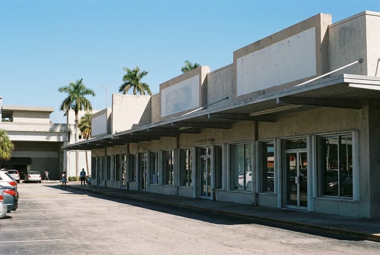 A low row of shopfronts fronting a wide car park under strong sun, flat canopy overhang and blank sign panels above the units