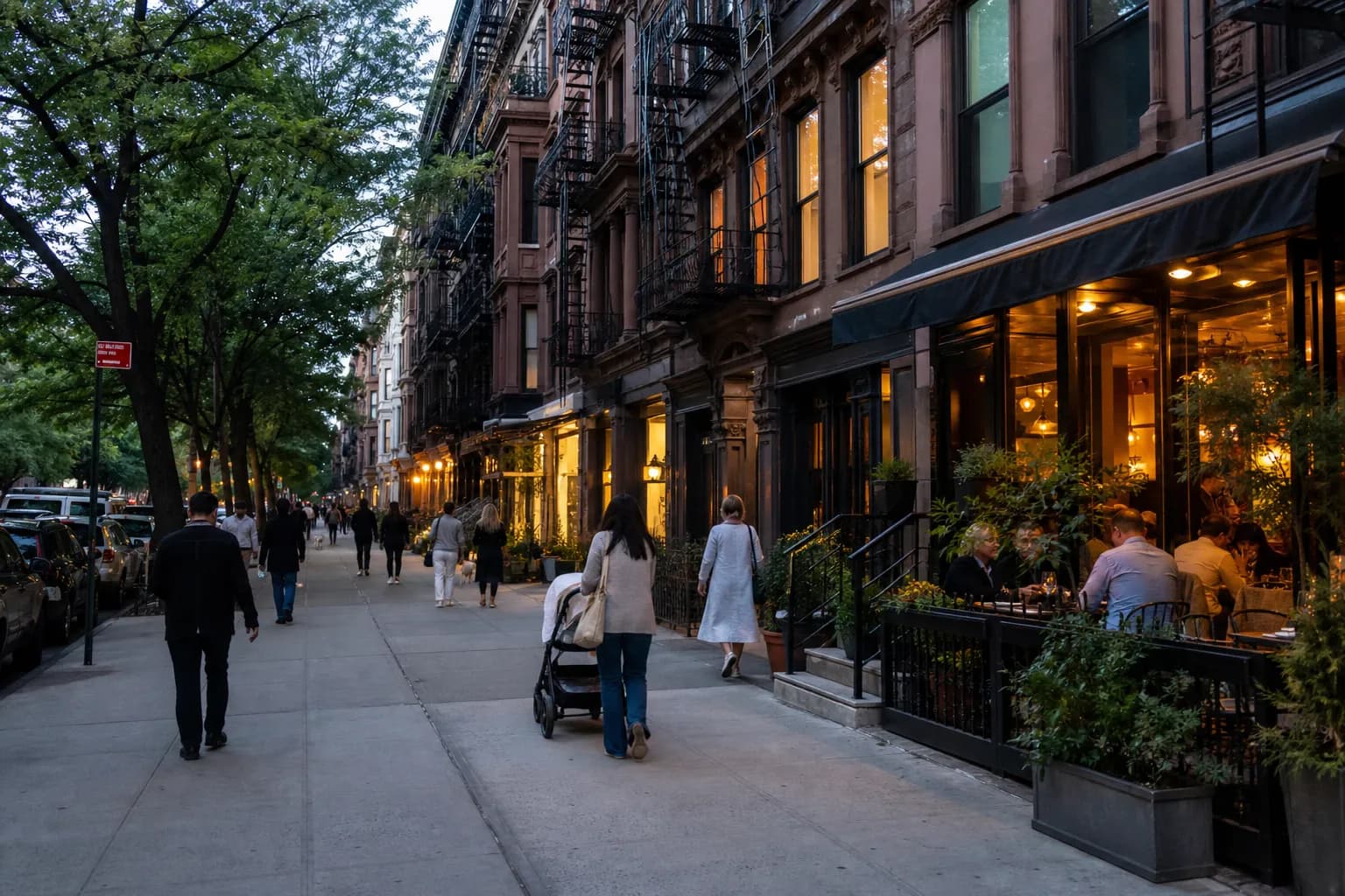 Tree-lined Upper West Side block with warm restaurant windows and a stroller on the sidewalk