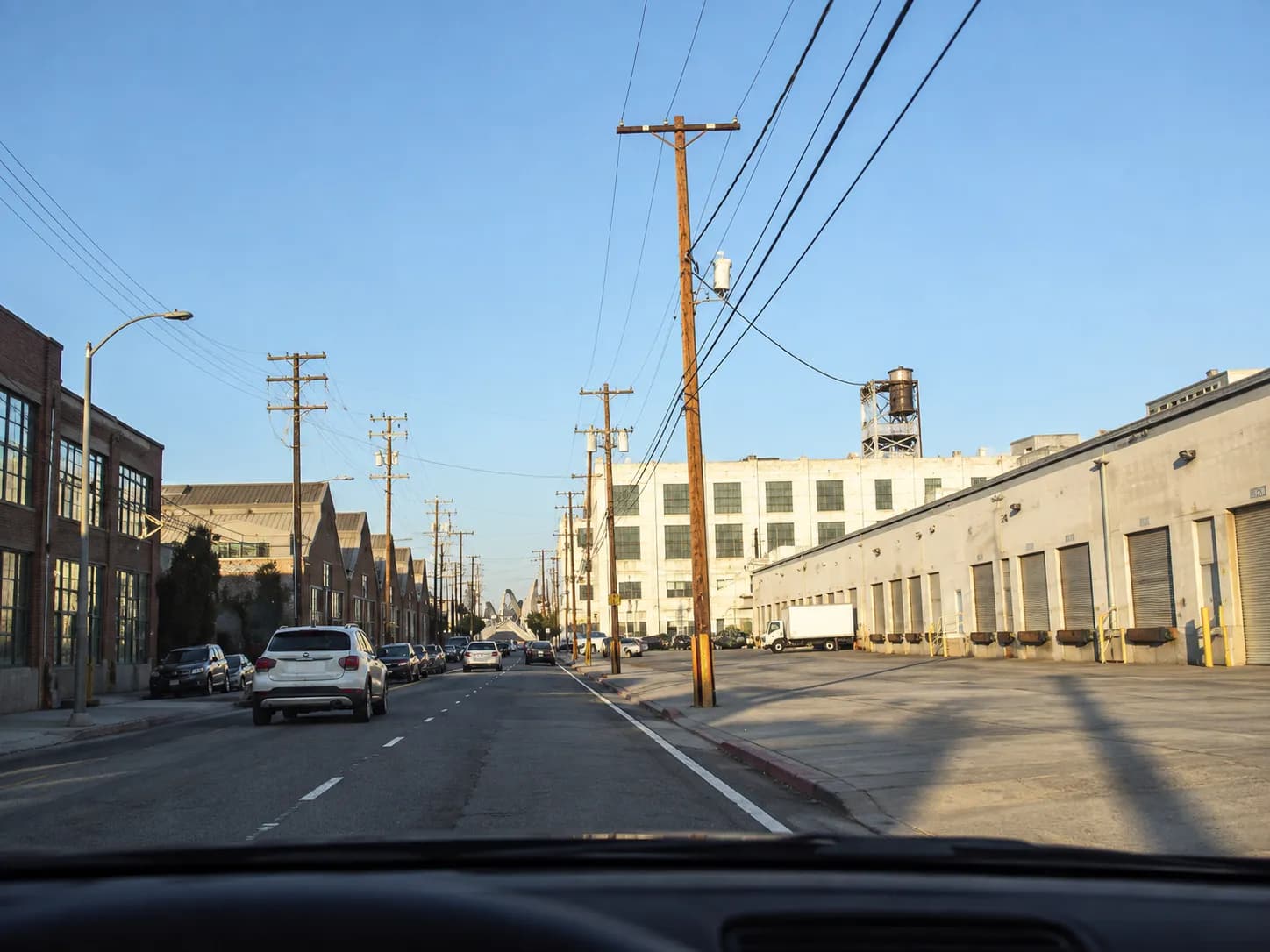 Industrial warehouse street with brick and concrete loading facades, roller shutters and power lines overhead under hard afternoon sun