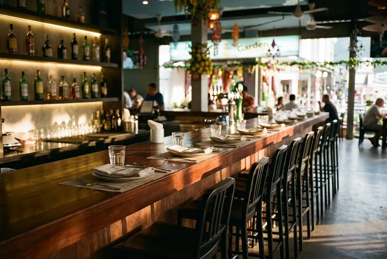 A polished bar counter with a row of high stools and place settings laid along it, bottles softly lit on the back shelf