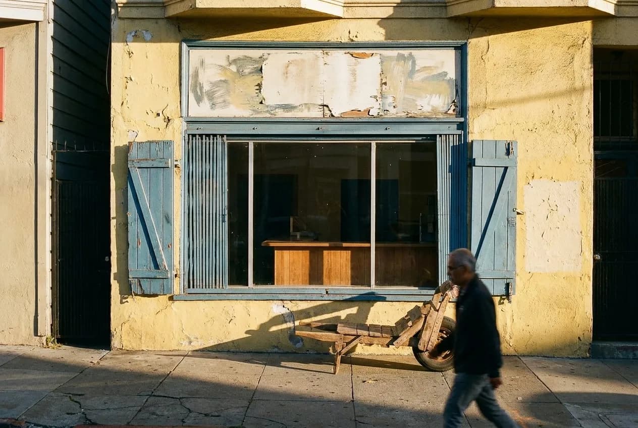 A narrow shopfront with a counter visible through the glass and a hand-painted sign panel above, sunlight across the pavement in front