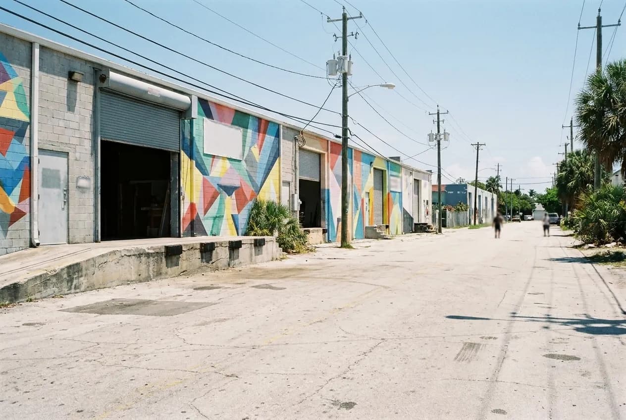 A wide street of low warehouse frontages with painted flanks under hard midday sun, roller doors and a kerbside loading bay in the foreground