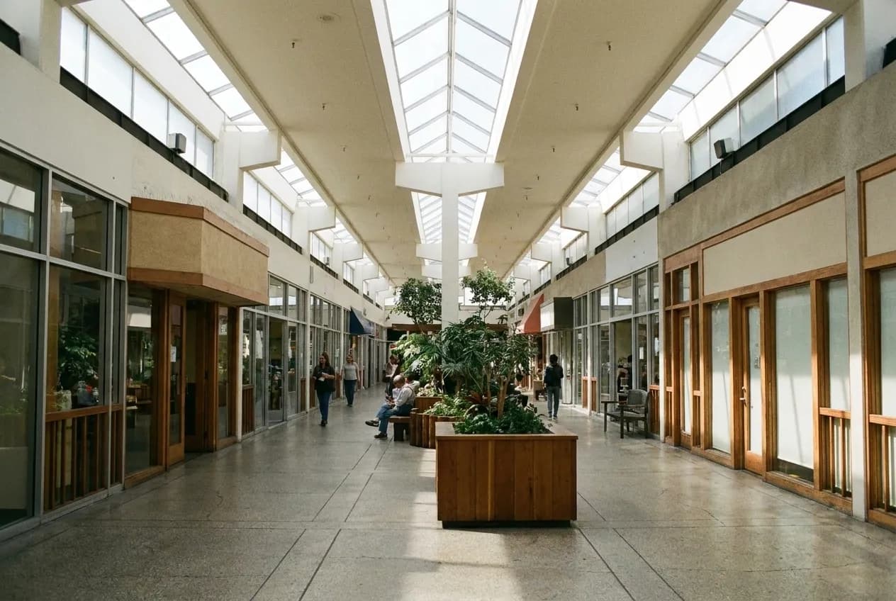 A covered indoor arcade with shopfronts along both sides and a skylit ceiling, planters down the middle and a tiled floor receding into the distance