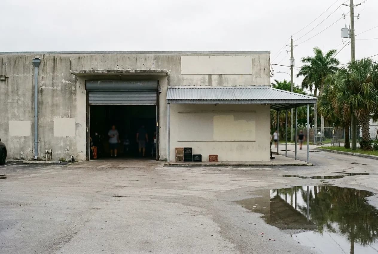 A low concrete warehouse frontage with a roller door standing open and a small awning beside it, painted render and a wide apron of tarmac in front