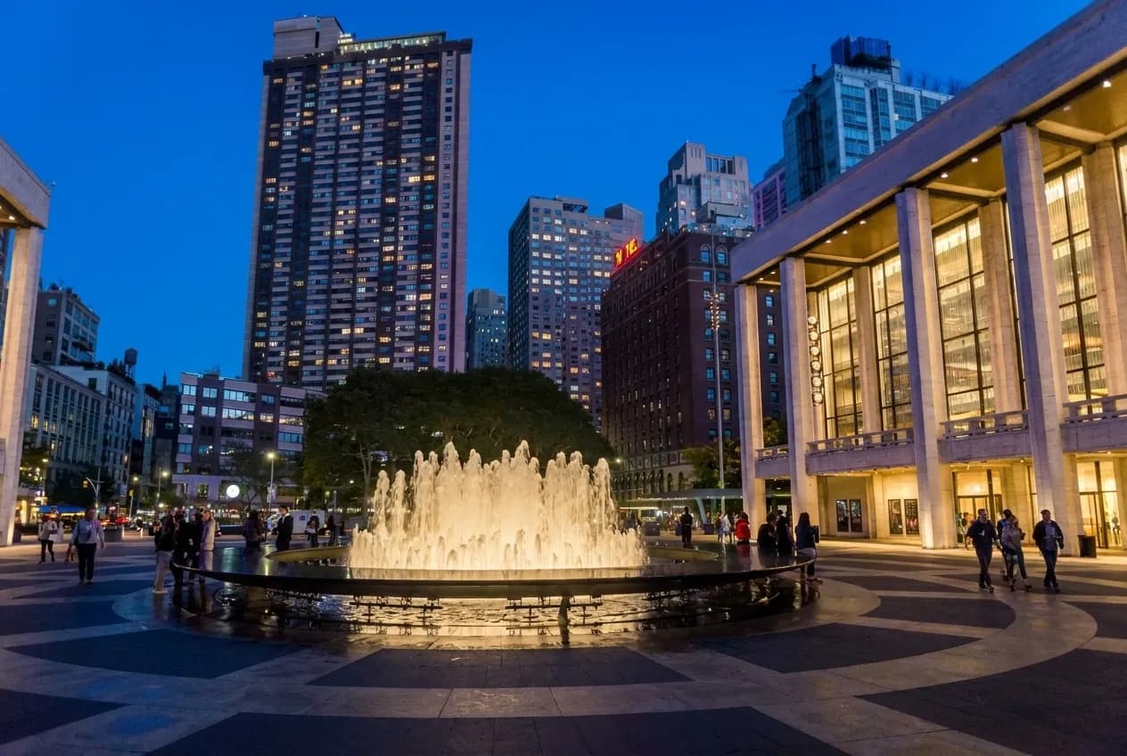 Wide dusk view of Lincoln Center's plaza with the illuminated Revson Fountain and the colonnade of David Geffen Hall