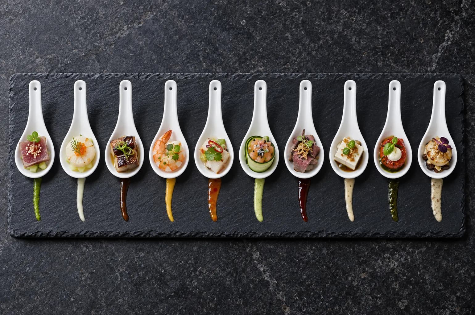 Row of small porcelain spoons on a slate tray, each holding a single precise bite with a thin sauce line beneath