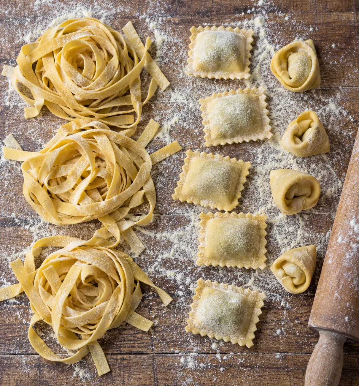 Fresh pasta ribbons and filled shapes laid in rows on a floured wooden board with a rolling pin at the edge