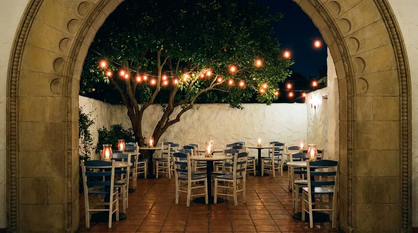 A candlelit courtyard restaurant at night with small tables under a tree, blue-and-white chairs and low garden lighting