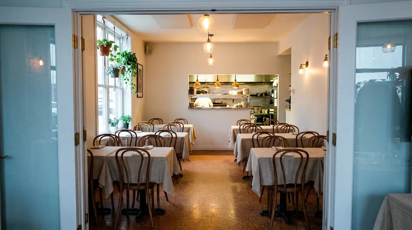 A small dining room seen through an open doorway, closely spaced tables set with white cloth, a pass at the back lit brighter than the room and hanging plants at the window