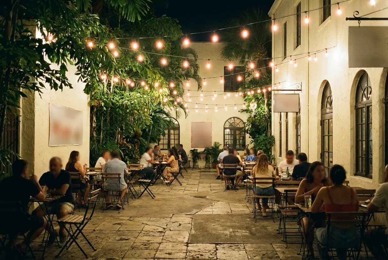 A small paved courtyard set with tables under strung lights and overhanging planting, warm pools of light and pale render walls on three sides