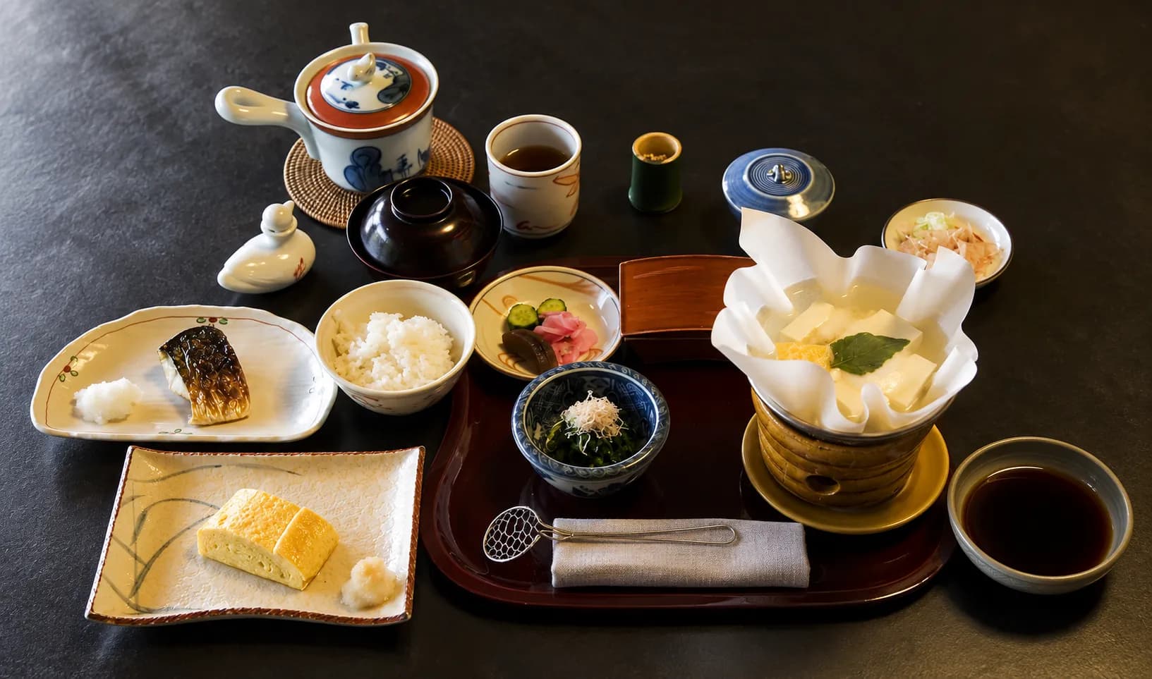 Small lacquer bowl and two ceramic dishes on a dark tray holding a seasonal arrangement of fish and vegetables