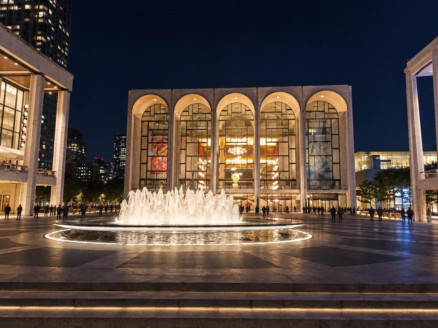 Modernist performing arts plaza at night with travertine halls around a circular fountain and glowing arched glass facades