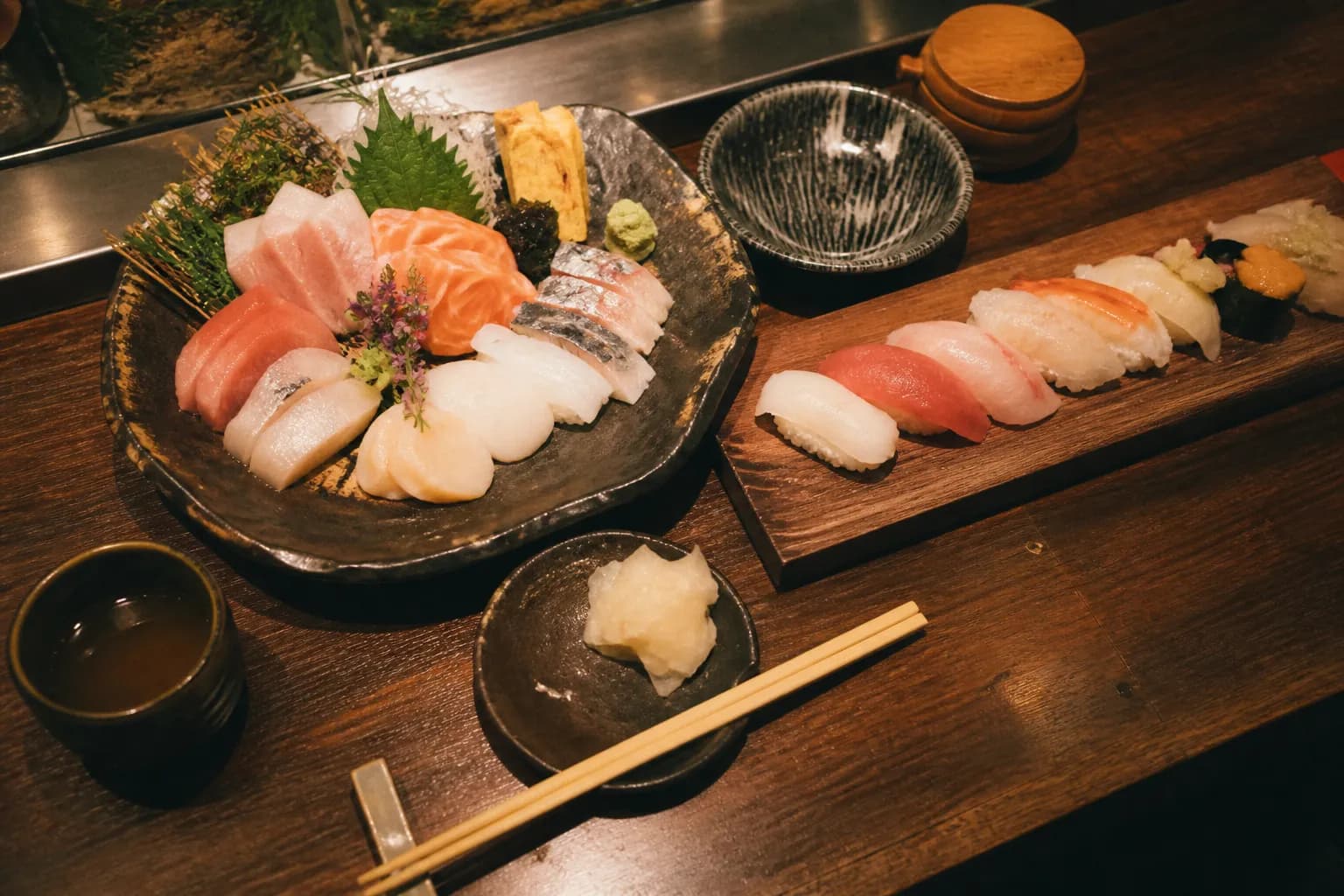 Overhead spread of an assorted sushi platter and fresh nigiri on a dark wooden counter with ginger and chopsticks in warm light