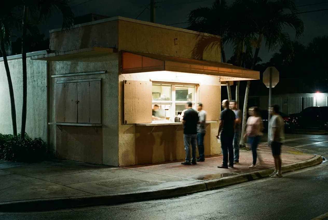 A small lit food counter open onto a dark street after midnight, warm light spilling onto the kerb and a few figures blurred in the queue
