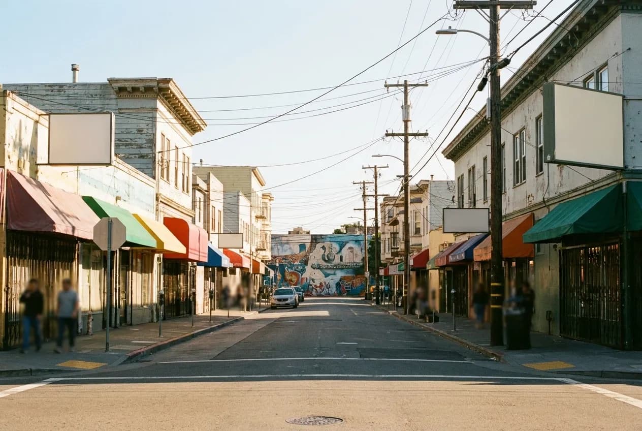 A wide sunlit street of low commercial buildings with a painted wall at one end, awnings over the pavement and utility wires crossing above