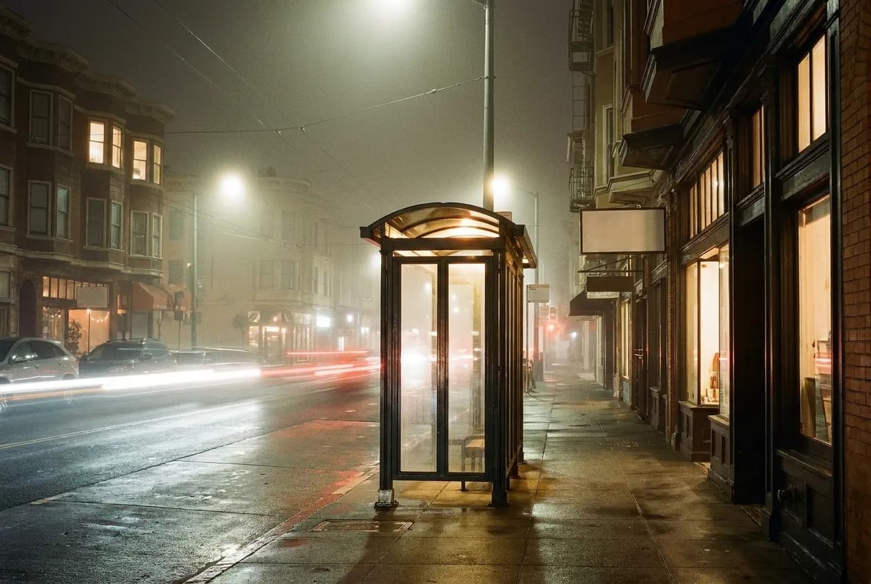 A late-night city street with a lit transit shelter and empty pavement, headlights streaking past and fog softening the shopfront glow behind
