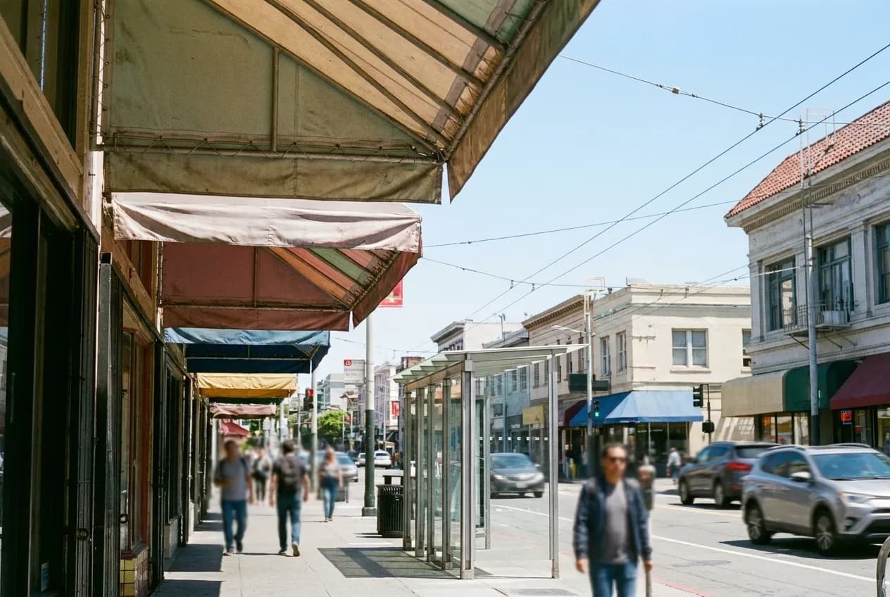 A busy commercial street with layered shop awnings and hand-painted frontages under bright sun, a bus stop shelter in the middle distance