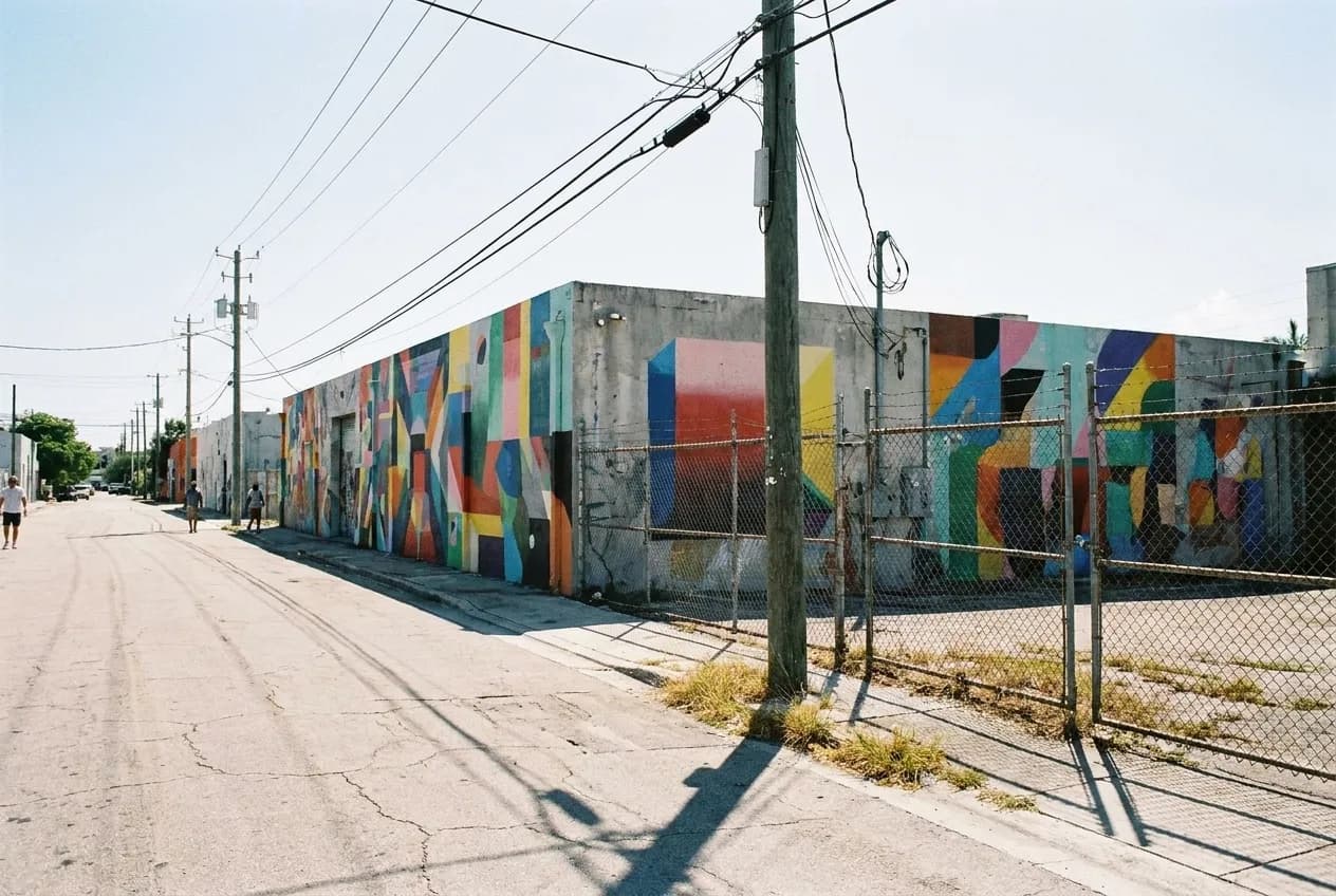 A low warehouse wall covered edge to edge in bright painted colour on a wide Miami street, chain-link and a utility pole in the foreground under strong sun