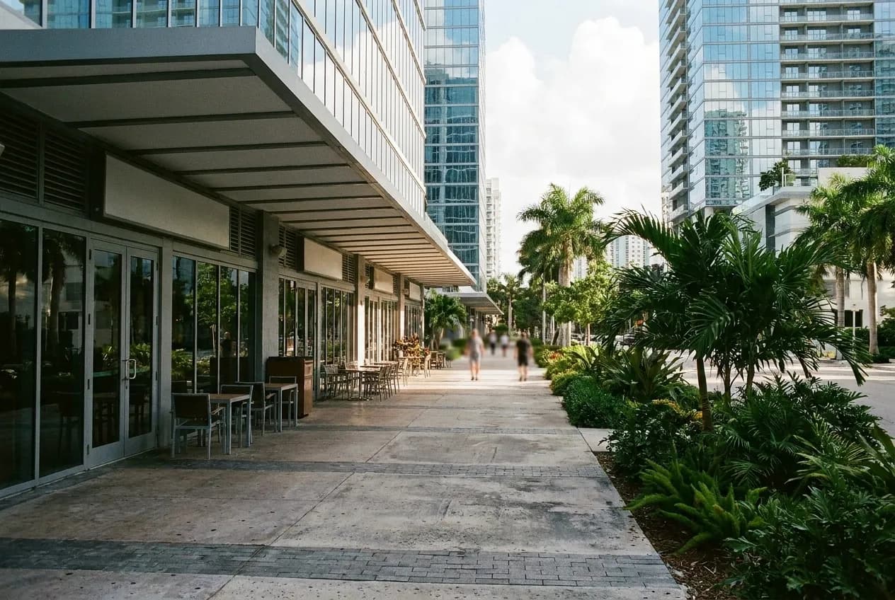 A covered walkway at the base of glass towers with restaurant frontages along one side, planted beds and a wide paved surface underfoot