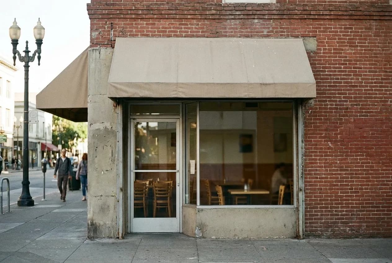 A modest restaurant frontage on a city street with a plain awning and a glass door, tables just visible inside and the pavement empty in front