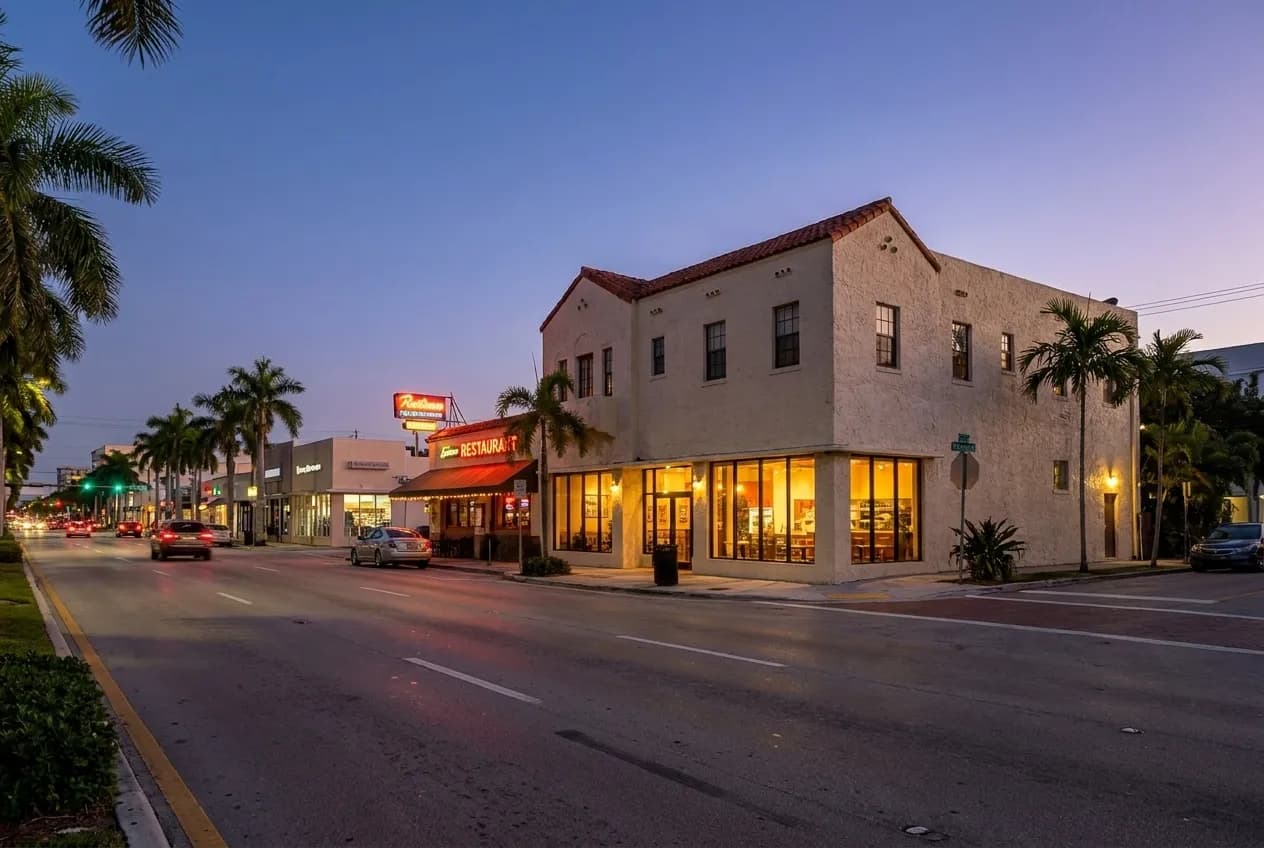 Wide arterial boulevard at dusk in Little River, low-rise storefronts glowing through plate glass, palm silhouettes against violet Miami sky, cars parked along Biscayne Boulevard.