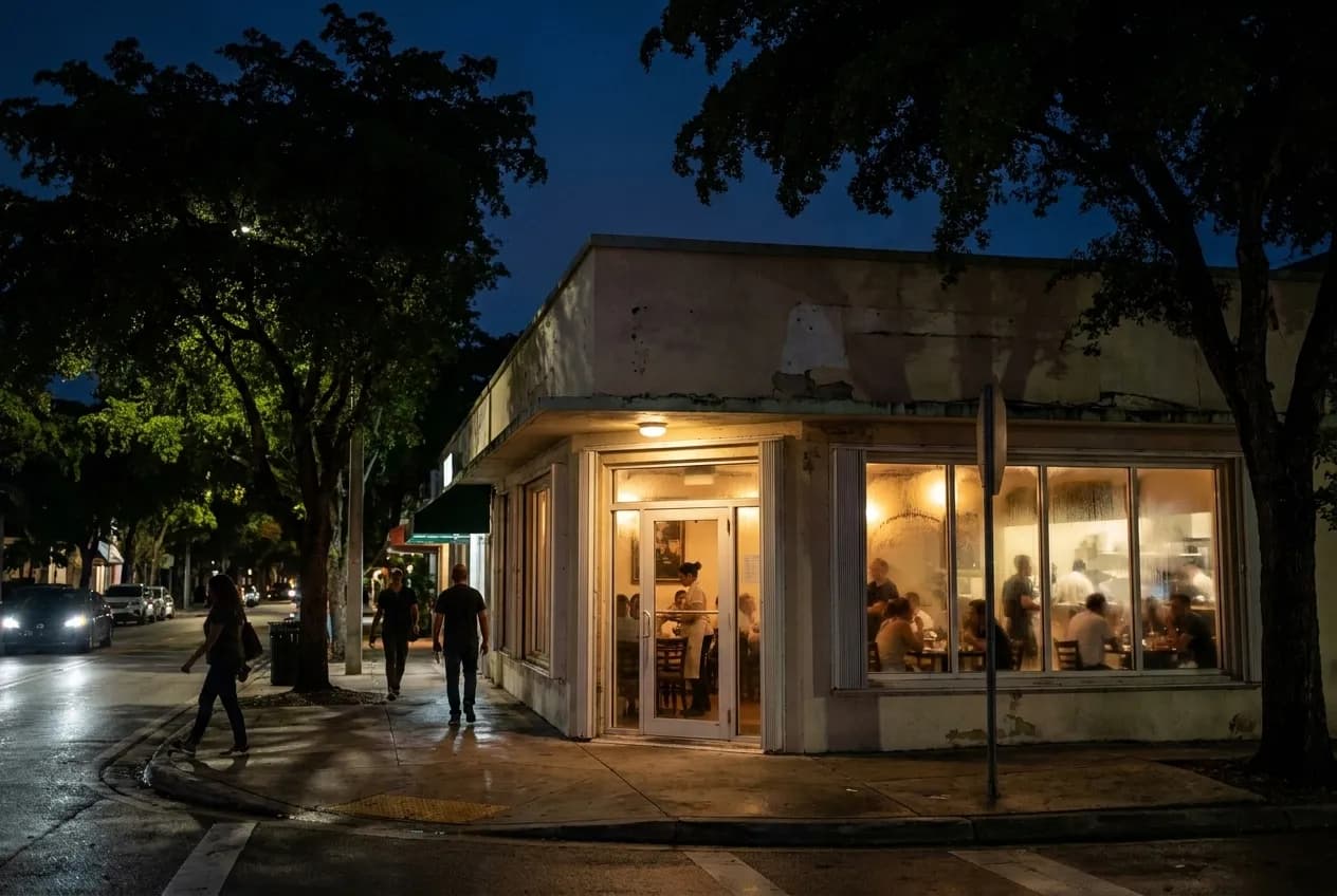 Evening glow on a leafy Coconut Grove corner restaurant with outdoor seating, warm interior light spilling onto the sidewalk and low-rise buildings visible beyond.