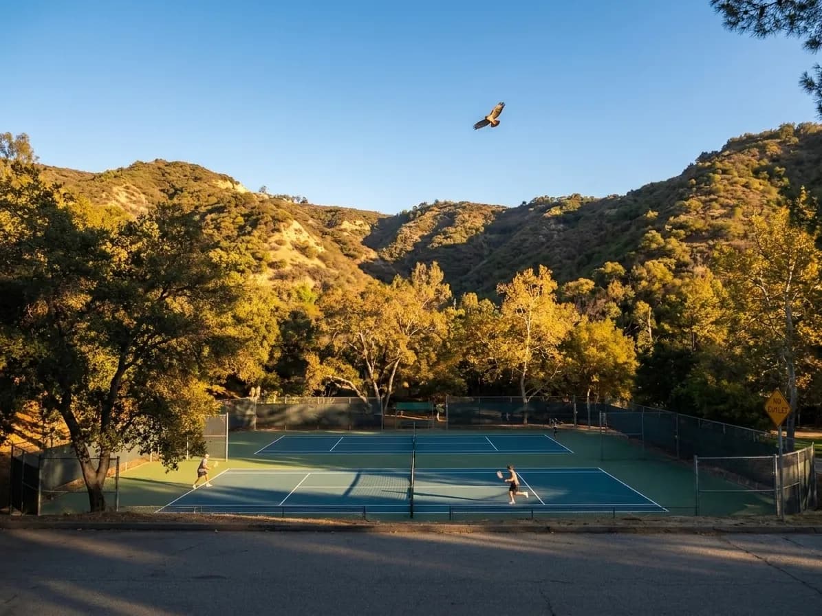 Two-tiered tennis courts nestled in a tree-lined canyon with chaparral hillsides rising behind the blue-green playing surfaces