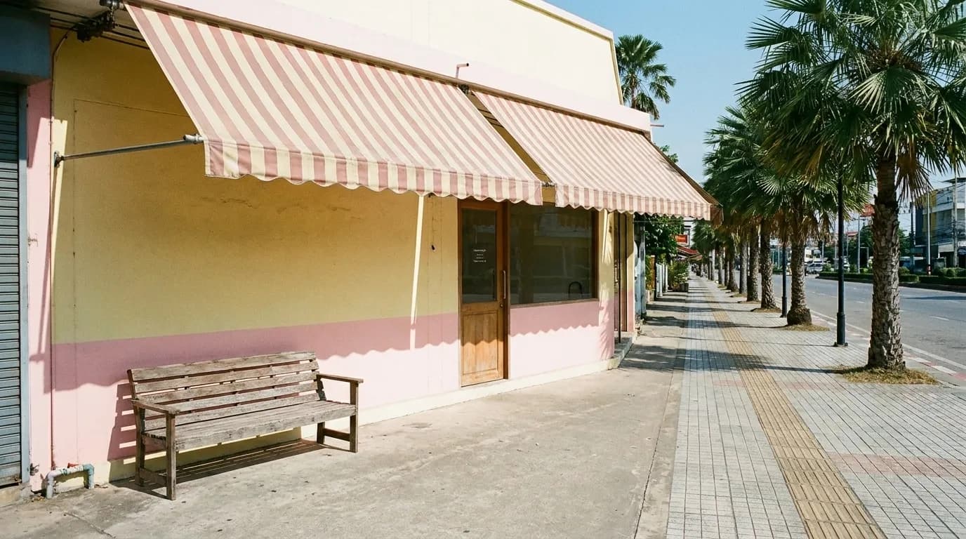A pastel-painted storefront on a wide sunlit street with awnings, a bench outside and palms along the pavement