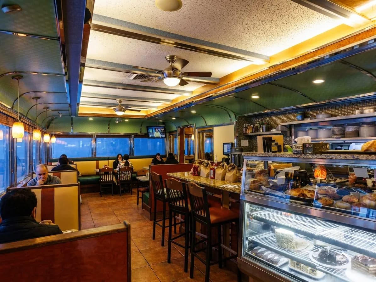 Railcar diner interior at dawn with counter stools, granite countertop, pastry case and booths under a curved green ceiling