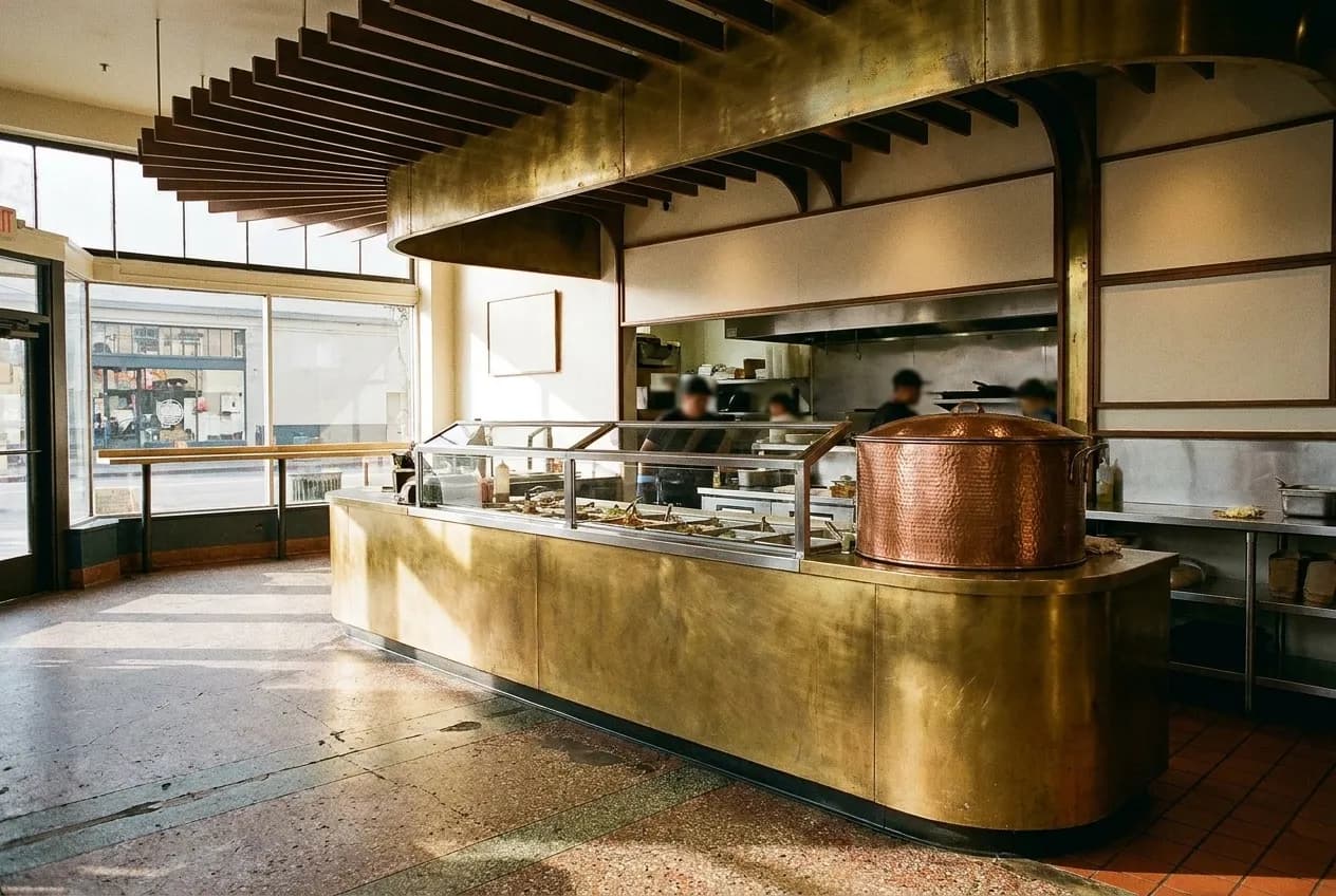 A taqueria counter with a steam table behind it and a tortilla warmer at one end, morning light from a shopfront window across the floor