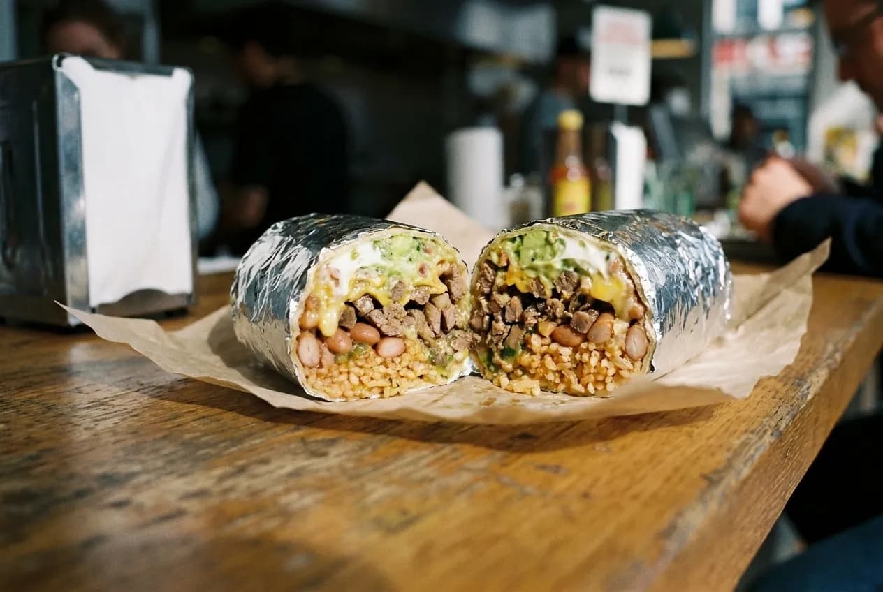 A large foil-wrapped burrito cut across the middle showing the layered filling inside, resting on paper on a counter