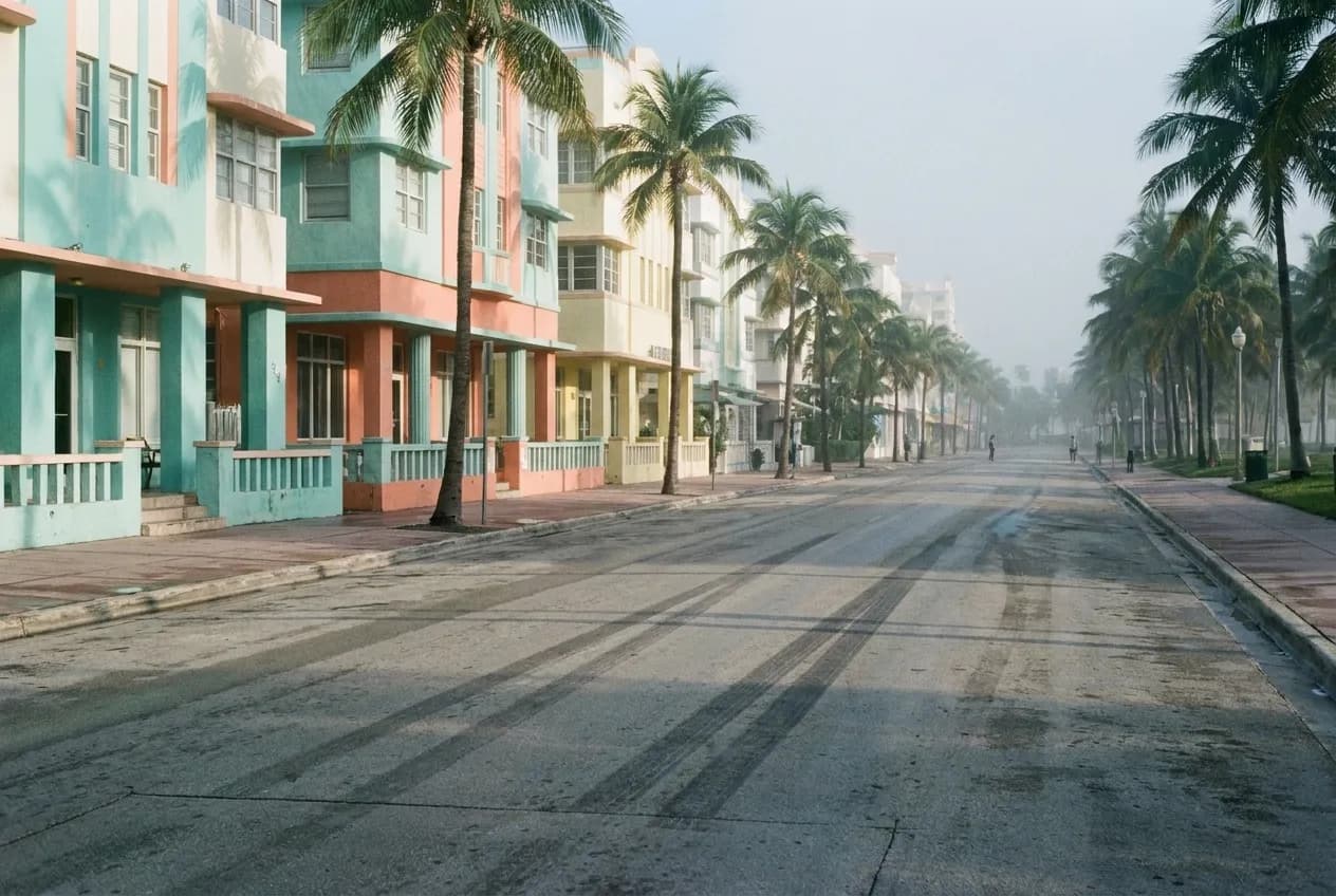 A quiet beachside street in early morning light with palms casting long shadows across empty pavement and low pastel buildings on one side