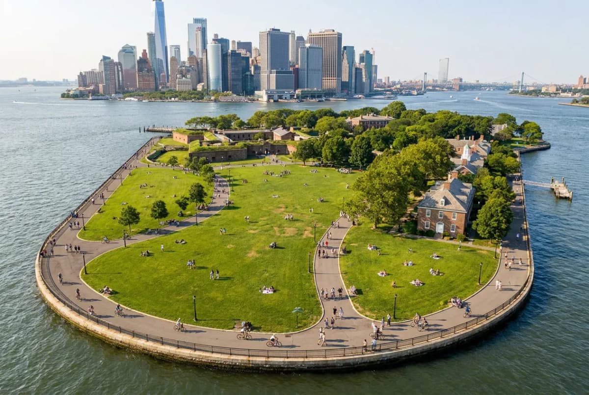 A green car-free island park with people walking and cycling on paths, the Manhattan skyline across the harbor in the distance