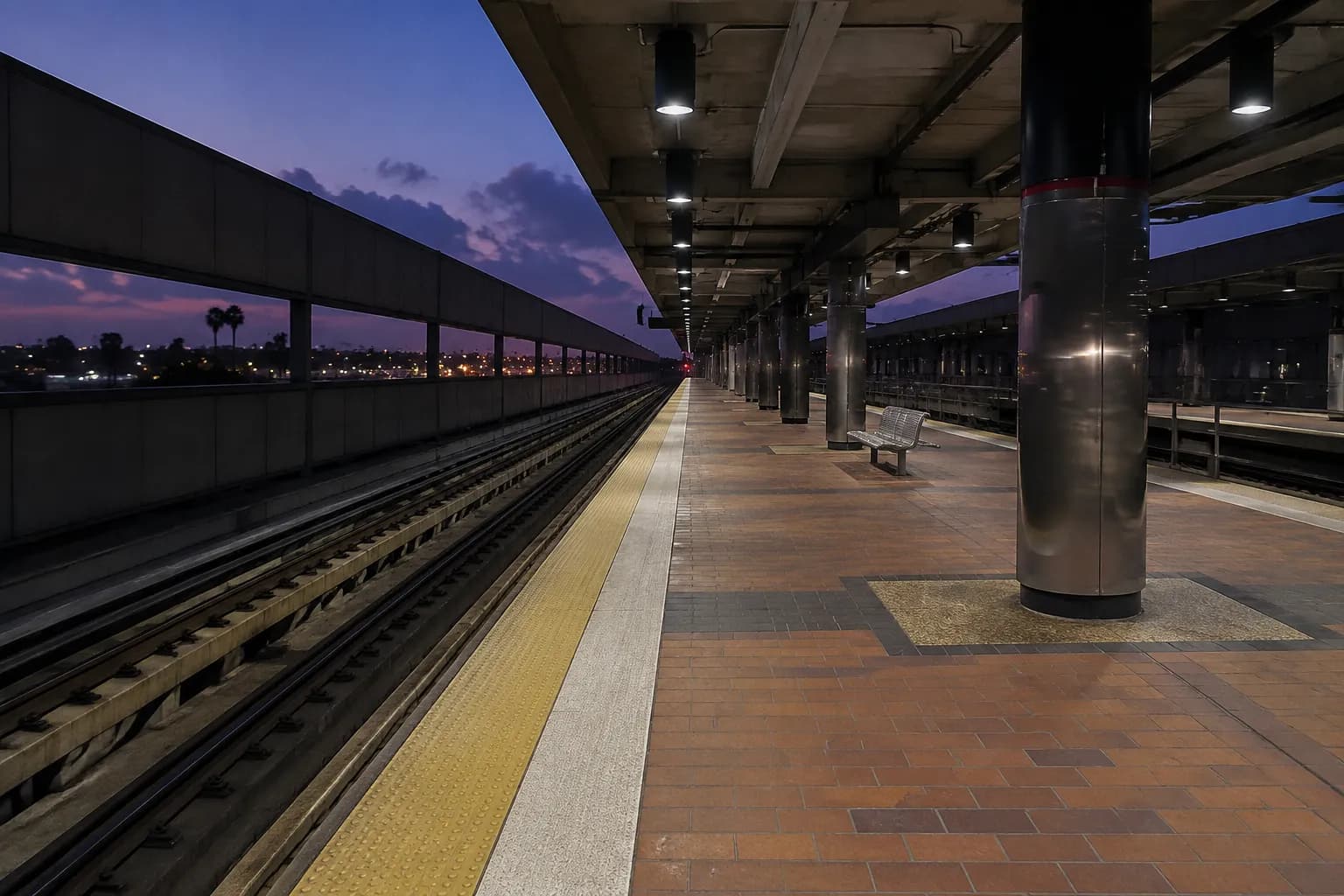 Metro E Line station platform at dusk with empty tracks and overhead lights beginning to illuminate