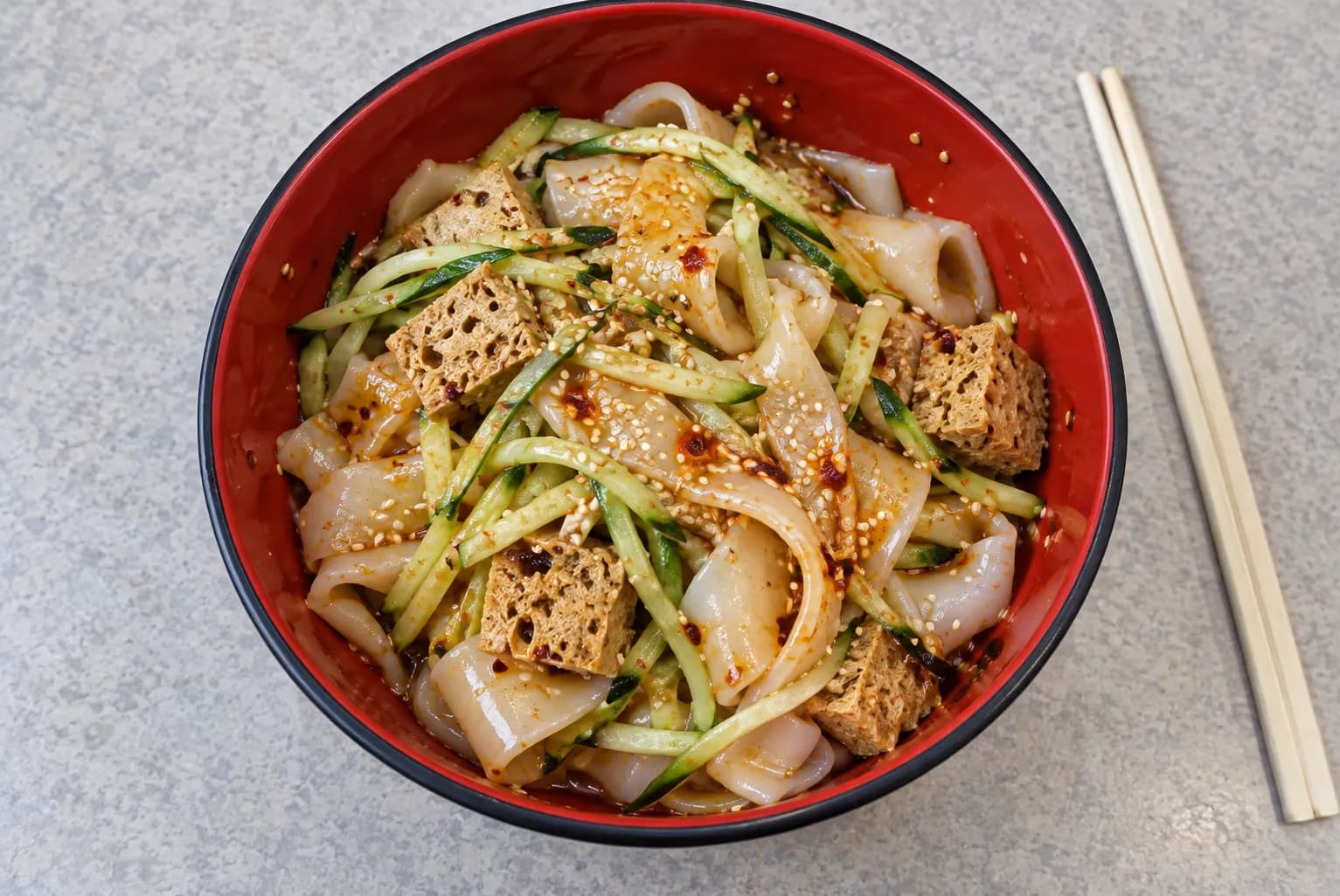 A cold noodle dish of wide translucent wheat-gluten strips in vinegar dressing with cucumber, in a plastic bowl