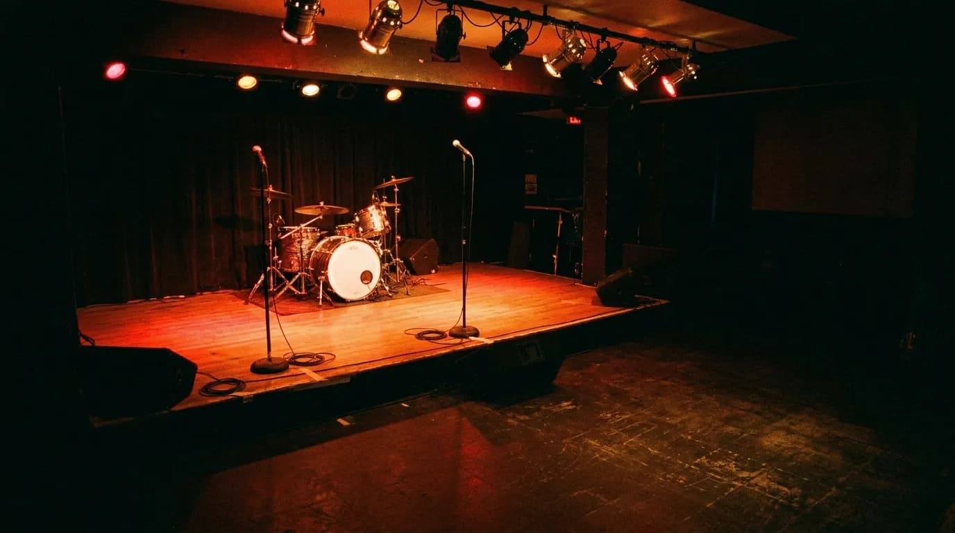 A small club stage lit in red with a drum kit and two microphone stands, seen from the back of an empty room