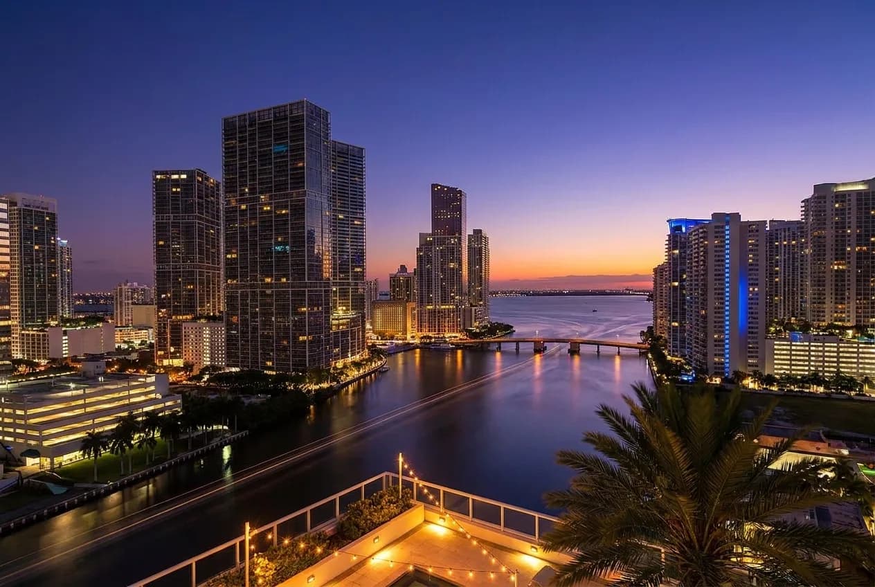 Dusk over the Brickell district of Miami seen from a high rooftop terrace, lit glass towers above a bay and bridge with a palm frond framing the corner