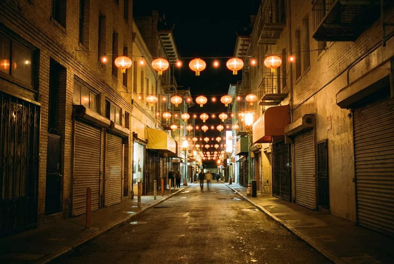 A narrow street after dark hung with lit lanterns, shopfront shutters mostly down and warm light pooling on the pavement beneath