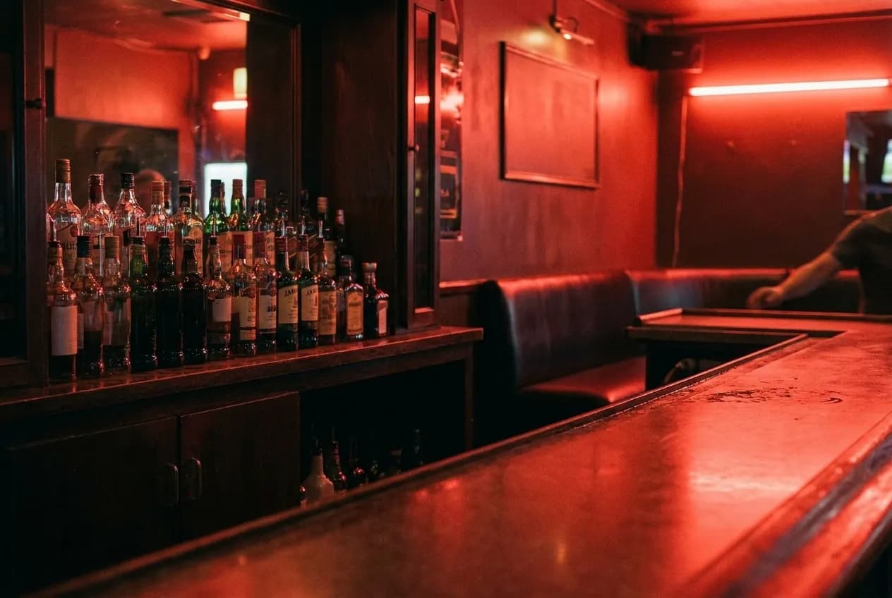 The interior of a dim bar washed in red light, a low booth against one wall and bottles ranked in shadow behind the counter