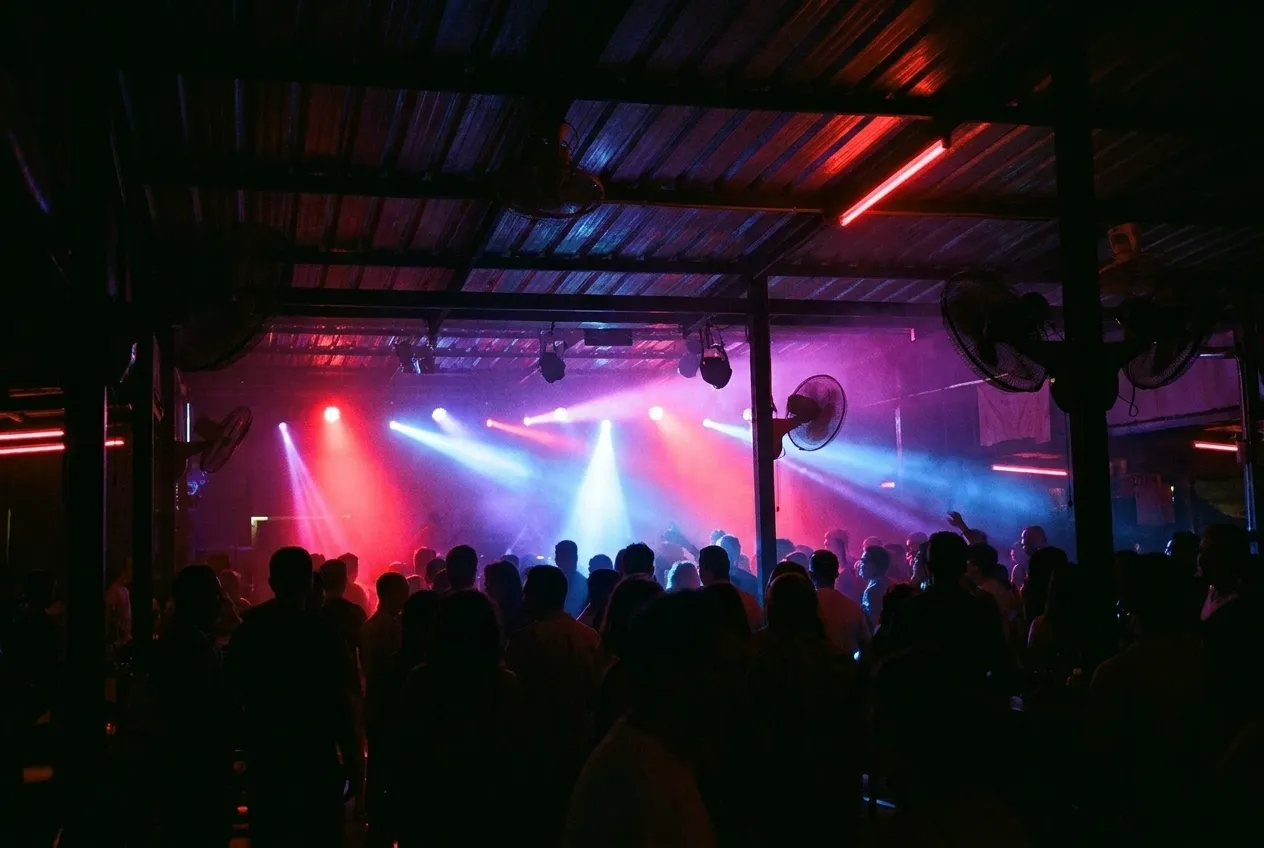A dark club interior with beams of coloured light cutting across a crowd, figures reduced to silhouettes against the glow of the stage end