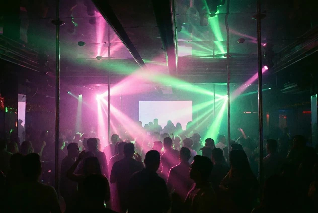 A dark interior with beams of coloured light over a crowded floor, figures reduced to silhouettes and haze catching the light above
