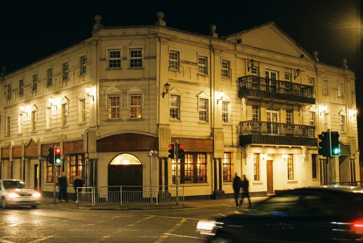 A corner bar building at night with a plain facade and warm light spilling from the door