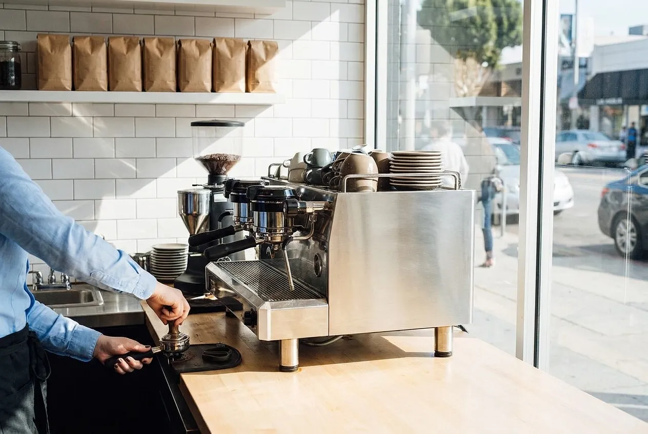 A bright Los Angeles coffee counter with a chrome espresso machine, stacked cups and hard daylight from a street-facing window